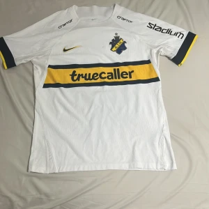 Signerad Vit AIK matchtröja L - AIK:s officiella vita matchtröja från Nike i Dri-FIT-material, storlek L Tröjan har korta ärmar, gula och svarta detaljer, klubbmärke på bröstet och sponsorer som Truecaller och SVEA. Nummer 20 och Sol Invictus på ryggen, samt flera autografer.