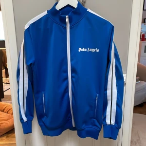 Blå träningsjacka från Palm Angels - Säljer en snygg blå träningsjacka från Palm Angels med vita ränder längs ärmarna och dragkedja framtill. Jackan har en hög krage och två fickor med dragkedja. Perfekt för en sportig look!