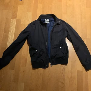 Svart bomberjacka från J.Lindeberg - Snygg svart bomberjacka från J.Lindeberg med dragkedja och ribbade muddar. Jackan har en stilren design med två fickor framtill och en diskret logga på bröstet. Perfekt för en cool och avslappnad stil.