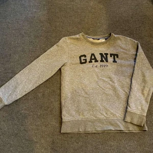 Grå sweatshirt från GANT - Säljer en stilren grå sweatshirt från GANT med svart text på framsidan. Tröjan har långa ärmar och en rund halsringning. Perfekt för en avslappnad look. Passar i alla tillfällen både i en skräm look och en stilren look.