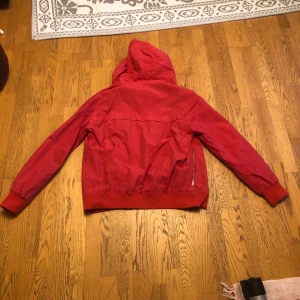 Röd vindjacka med huva - Säljer en snygg röd vindjacka, Perfekt för blåsiga dagar, fick ärva den av min pappa för kanske 2 eller 3 år sedan, men den har slutat passat mig nu