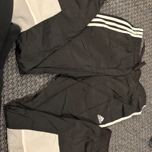Svarta träningsbyxor från Adidas - Säljer ett par svarta träningsbyxor från Adidas med vita ränder längs sidorna. Byxorna har en elastisk midja med snörning och är tillverkade i ett syntetmaterial. Perfekta för sport och träning.
