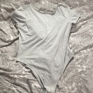 Grå bodysuit från H&M Divided - Säljer en enkel och stilren grå bodysuit från H&M Divided. Den har korta ärmar och en rund halsringning, perfekt för en avslappnad look. Materialet är mjukt och stretchigt för en bekväm passform.