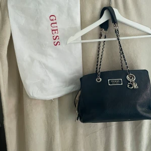 Svart axelväska från Guess - Snygg svart axelväska från Guess med kedjedetaljer och logotypmönster. Väskan har en dragkedja och flera fack inuti för enkel organisering. Perfekt för att ge din outfit en stilfull touch. Bekväm och får plats med mycket. 
