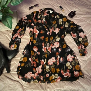 Blommig klänning med knappar - Säljer en svart klänning med färgglatt blommönster i rosa, orange och gult. Klänningen har långa ärmar och knappar framtill. Perfekt för en stilfull look, eller som en fin cardigan på stranden! Superfin och knappt använd men en av knapparna på ärmarna går inte att knäppa 