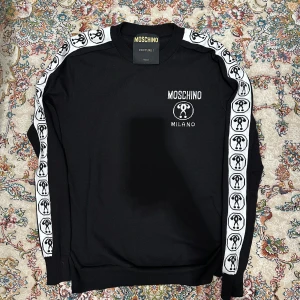 Moschino tröja - Snygg svart tröja från Moschino med vita detaljer och logotyp på bröstet. Tröjan har långa ärmar med ett mönstrat band längs ärmarna. Perfekt för en stilren look.
