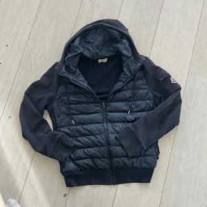 Svart/Mörkblå  cardigan från Moncler - Snygg cardigan jacka från Moncler med huva och dragkedja. Jackan har en quiltad design på framsidan och ärmar i ett annat material. Perfekt för en stilren look.