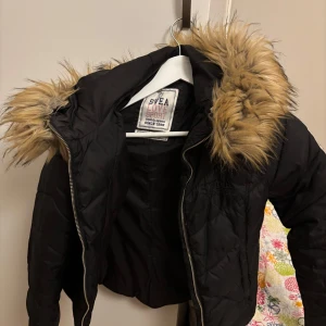 Svart jacka med päls från Svea - Snygg svart jacka från Svea med quiltad design och en mysig pälsfodrad huva. Jackan har dragkedja framtill och en broderad logga på ärmen. Perfekt för kyliga dagar! Fickorna går inte att stänga. Passar 10-12 år