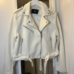 Vit jacka från BikBok - Snygg vit jacka från BikBok med dragkedjor och bälte i midjan. Jackan har en klassisk bikerstil med långa ärmar och silverfärgade detaljer. Perfekt för en stilren look.