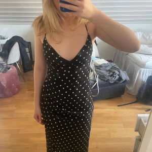 Svart prickig klänning med öppen rygg - Snygg svart klänning med vita prickar och en elegant öppen rygg med snörning. Klänningen har smala axelband och en figurnära passform som ger en stilren look. Perfekt för sommarens fester!