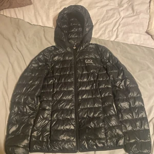 Svart dunjacka från Emporio Armani - Säljer en snygg svart dunjacka från Emporio Armani EA7. Jackan har en quiltad design med dragkedja och huva. Perfekt för kyliga dagar och ger en stilren look.