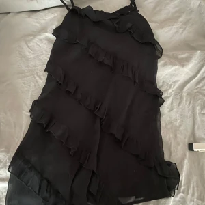 Svart klänning från Gina Tricot - Snygg svart klänning från Gina Tricot med volangdetaljer. Klänningen har smala axelband och en asymmetrisk design som ger en trendig look. Perfekt för en kväll ute eller en speciell tillställning. Använd två gånger och köpt för 200kr