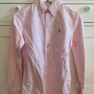 Rosa randig skjorta från Ralph Lauren - Säljer en stilren rosa och vit randig skjorta från Ralph Lauren. Skjortan har långa ärmar och knappar framtill. Den är i en custom fit och har det klassiska Ralph Lauren-logotypen broderad på bröstet. Perfekt för en snygg och avslappnad look.