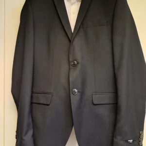 Svart kostym från Jack & Jones - Snygg svart kostym med vit skjorta från Jack & Jones i super slim passform. Kavajen har två knappar och klassiska fickor. Byxorna är stilrena och matchar perfekt med kavajen. Perfekt för skolavslutning eller student. Storlek 46 på kavajen och 44 på byxorna. Skicka meddelande för fler bilder. 