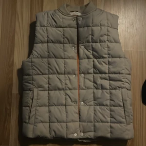 Beige quiltad väst - Säljer en stilren beige quiltad väst med knappar framtill. Västen har en klassisk design med en bekväm passform och ribbad krage. Perfekt för lager-på-lager under kyligare dagar.
