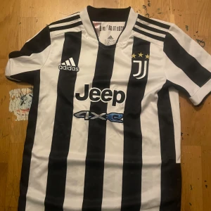 Juventus fotbollströja från Adidas - Säljer en svartvit randig fotbollströja från Adidas med korta ärmar. Tröjan har Juventus-logga och Jeep-sponsormärke på framsidan. Perfekt för fotbollsfans som vill visa sitt stöd för laget. Tillverkad i Primegreen-material. Storleken är 11-12 år/152cm. Köptes år 2021 för 900kr på unisport.