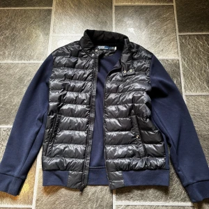 Polo Ralph lauren cardigan - Snygg blå jacka från Ralph Lauren med quiltad front och dragkedja. Jackan har långa ärmar och en liten broderad logga på ärmen. Perfekt för en stilren look. Passar ca 180-185cm
