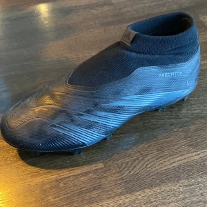 Svarta Adidas Predator fotbollsskor - Säljer ett par svarta Adidas Predator fotbollsskor. Skorna har en strumpliknande passform utan snörning och är designade för att ge bra grepp på planen. Perfekta för den seriösa fotbollsspelaren som vill ha kontroll och precision.
