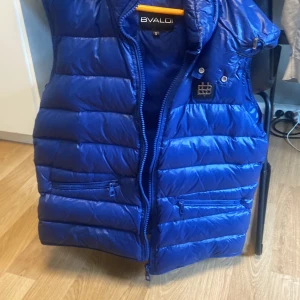 Blå dunväst från Bvaldi - Snygg blå dunväst från Bvaldi med dragkedja och praktiska fickor. Perfekt för kyliga dagar när du vill hålla stilen. Västen har en hög krage och en modern look.