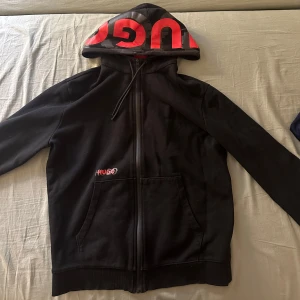 Svart hoodie från Hugo Boss - Säljer en svart hoodie från Hugo Boss med röd logga på huvan och bröstet. Tröjan har dragkedja och långa ärmar, perfekt för en casual look. Den har också praktiska fickor framtill.