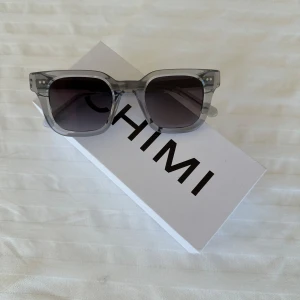Grå solglasögon från Chimi - Snygga grå solglasögon från Chimi med en fyrkantig form och transparent båge. Perfekta för att ge en cool touch till din stil. De mörka linserna ger ett bra skydd mot solen.