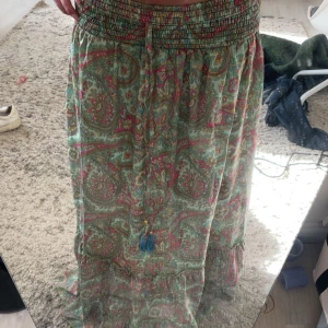 Mönstrad långkjol med volang - Säljer en färgglad långkjol med paisleymönster i grönt, rosa och beige. Kjolen har en elastisk midja och dekorativa tofsar framtill. Perfekt för en bohemisk look!