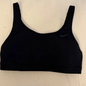 Svart sport-bh från Nike - Säljer en svart sport-bh från Nike med Dri-FIT-teknologi. Den har justerbara axelband och en bekväm passform. Perfekt för träning och ger bra stöd. Nike-loggan syns diskret på framsidan. Den är ovadderad. 