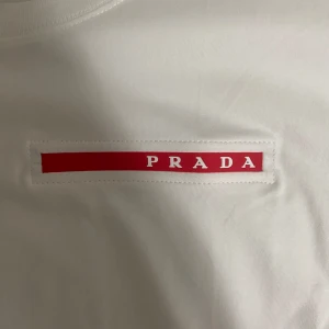 Vit t-shirt från Prada - Säljer en stilren vit t-shirt från Prada med en röd logga på bröstet. T-shirten har en klassisk passform och korta ärmar. Perfekt för en minimalistisk look.