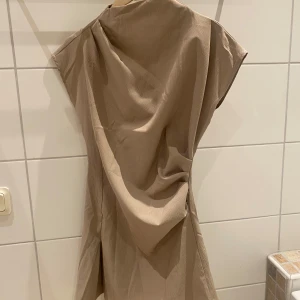 Beige klänning med korta ärmar - Snygg beige klänning med korta ärmar och en elegant drapering. Perfekt för en stilren look. Klänningen har en diskret struktur i tyget som ger en extra touch.