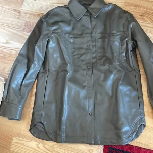 Olivgrön skinnskjorta - Snygg olivgrön skinnskjorta med knappar och en bröstficka. Perfekt för en stilren look med en touch av edge. Skjortan har långa ärmar och en klassisk krage.