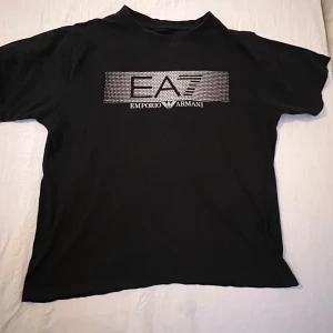 Svart t-shirt från Emporio Armani - Snygg svart t-shirt från Emporio Armani med ett stilrent EA7-tryck på bröstet. Perfekt för en casual look med en touch av lyx. Passar bra till jeans eller shorts för en avslappnad stil.