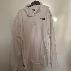 Vit fleece tröja från The North Face - Säljer en vit fleece tröja från The North Face med halv dragkedja och broderad logga på bröstet. Perfekt för kyliga dagar med sin mjuka och varma känsla. Tröjan har långa ärmar och en hög krage för extra värme.