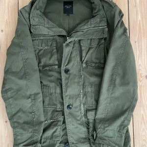 Safari jacka från New Look Men - Säljer en grön safari jacka från New Look Men i storlek S. Jackan har en dragkedja och knappar framtill samt flera praktiska fickor och några slitningar som ger en unik stil. Perfekt för lite kallare vår klimat. Nypris 1800!
