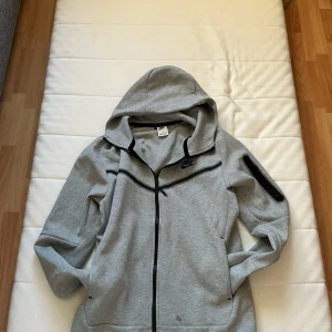 Grå Nike Tech - Säljer en grå huvtröja från Nike med dragkedja och svart logotyp på bröstet. Tröjan har en praktisk ficka på ärmen. Perfekt för en sportig look eller avslappnade dagar.