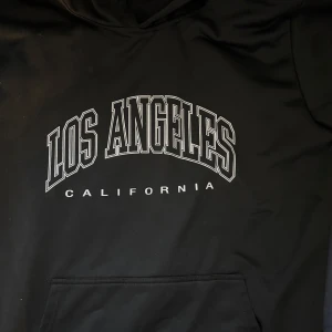 Svart hoodie med Los Angeles-tryck - Säljer en svart hoodie med stort tryck av 'Los Angeles California' på framsidan. Perfekt för en avslappnad stil med känguruficka framtill. Passar bra för kyliga dagar.