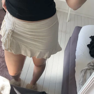 Beige kjol från Stradivarius - Säljer en söt beige kjol från Stradivarius med volangdetaljer och knytning på sidan. Kjolen har en dragkedja för enkel påklädning och är perfekt för varma dagar. Den har även insydda shorts. 
