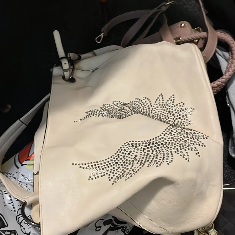 Snygg vit axelväska med dekorativa vingdetaljer i glittrande stenar på framsidan. Väskan har flätade handtag och en justerbar axelrem. Perfekt för att ge en stilfull touch till din outfit.. Laukut & Käsilaukut.
