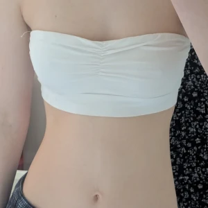 Vit tubtopp från Gina Tricot - Säljer en stilren vit banduetopp från Gina Tricot. Toppen är ärmlös och har en rynkad detalj framtill som ger en snygg touch. Perfekt för varma sommardagar, istället för bh eller som ett basplagg i garderoben.