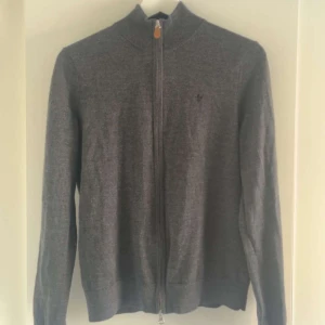 Grå merinoulltröja från Morris - Säljer en stilren grå tröja från Morris i 100% merinoull. Tröjan har en dragkedja framtill och långa ärmar, perfekt för kyligare dagar. Den har en liten broderad logga på bröstet. Passar både till vardag och mer uppklädda tillfällen.