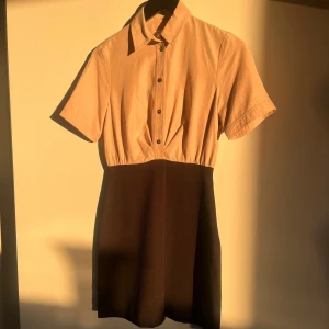 Beige och svart klänning från Sandro - Snygg klänning från Sandro i beige och svart. Den har korta ärmar och knappar framtill. Perfekt för en stilren look.