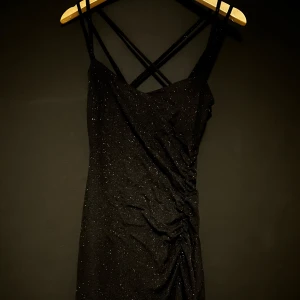 Svart glittrig klänning med smala axelband - Elegant svart kort klänning med glittrande detaljer och smala axelband som korsas i ryggen. Perfekt för en kväll ute med en stilren och sofistikerad look. Är storlek XS men kan passa i S, har används en gång.