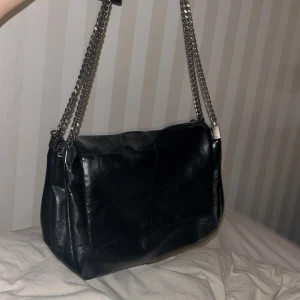 Zara Svart axelväska med kedjedetaljer - Snygg svart axelväska i skinn med stiliga kedjedetaljer på axelremmen. Väskan har en klassisk design med en praktisk storlek för vardagsbruk. Perfekt för att ge din outfit en edgy touch.