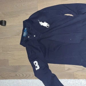 Mörkblå pikétröja från Ralph Lauren - Säljer en stilren mörkblå pikétröja från Ralph Lauren med klassisk krage och knappar. Tröjan har långa ärmar och ett stort vitt logotryck på bröstet samt siffran 3 på ärmen. Perfekt för en avslappnad men snygg look.