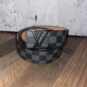 Rutig bälte från Louis Vuitton - Snyggt bälte från Louis Vuitton med ett klassiskt rutigt mönster i svart och grått. Bältet har en stilren spänne med LV-logotypen och är tillverkat i skinn. Perfekt för att ge din outfit en lyxig touch.