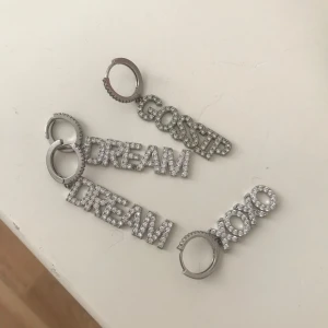 Örhängen med text i silver från IOAKU🥰🥰🫰🏻💞 - Snygga örhängen i silver med glittrande text. Varje örhänge har olika ord: 'GOSSIP', 'DREAM', och 'XOXO'. Perfekta för att ge en personlig touch till din stil.
