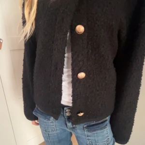 Svart cardigan jacka med guldknappar - Säljer en svart cardigan jacka med stora guldknappar framtill. Jackan har lång ärm och en mjuk, lite fluffig struktur. Passar perfekt till jeans för en avslappnad men snygg look.
