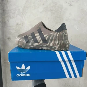 Adidas kamouflage slip-on skor - Unika slip-on skor från Adidas i kamouflagefärgat mönster med svarta ränder på sidan. Skorna har en rund tå och är tillverkade i syntetmaterial. Perfekta för dig som vill sticka ut med en modern och sportig look. Pris kan diskuteras 