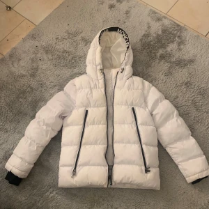 Vit dunjacka från Moncler - Säljer en vit dunjacka från Moncler med svart dragkedja och huva. Jackan har Moncler-logga på ärmen och svart textdetalj på insidan av huvan. Perfekt för kalla dagar och har en klassisk pufferstil.strlk 3 passar som M.Köpte den för 3000 andvänt få gånger 