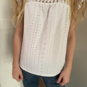 Vit blus med broderade detaljer - Säljer en söt vit blus med korta ärmar och broderade hålmönster. Blusen har en lös passform och är perfekt för varmare dagar. Den har fina spetsdetaljer upptill och är tillverkad i ett lätt bomullsmaterial.