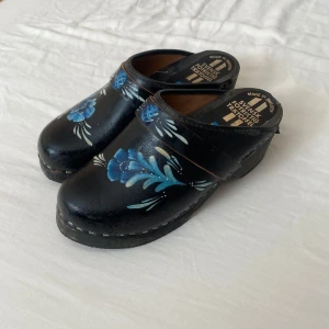 Svarta trätofflor med blått blommönster - Säljer ett par äkta svenska svarta trätofflor med handmålat blått blommönster. Klassisk svensk design med träbotten och öppen häl. I strl 35